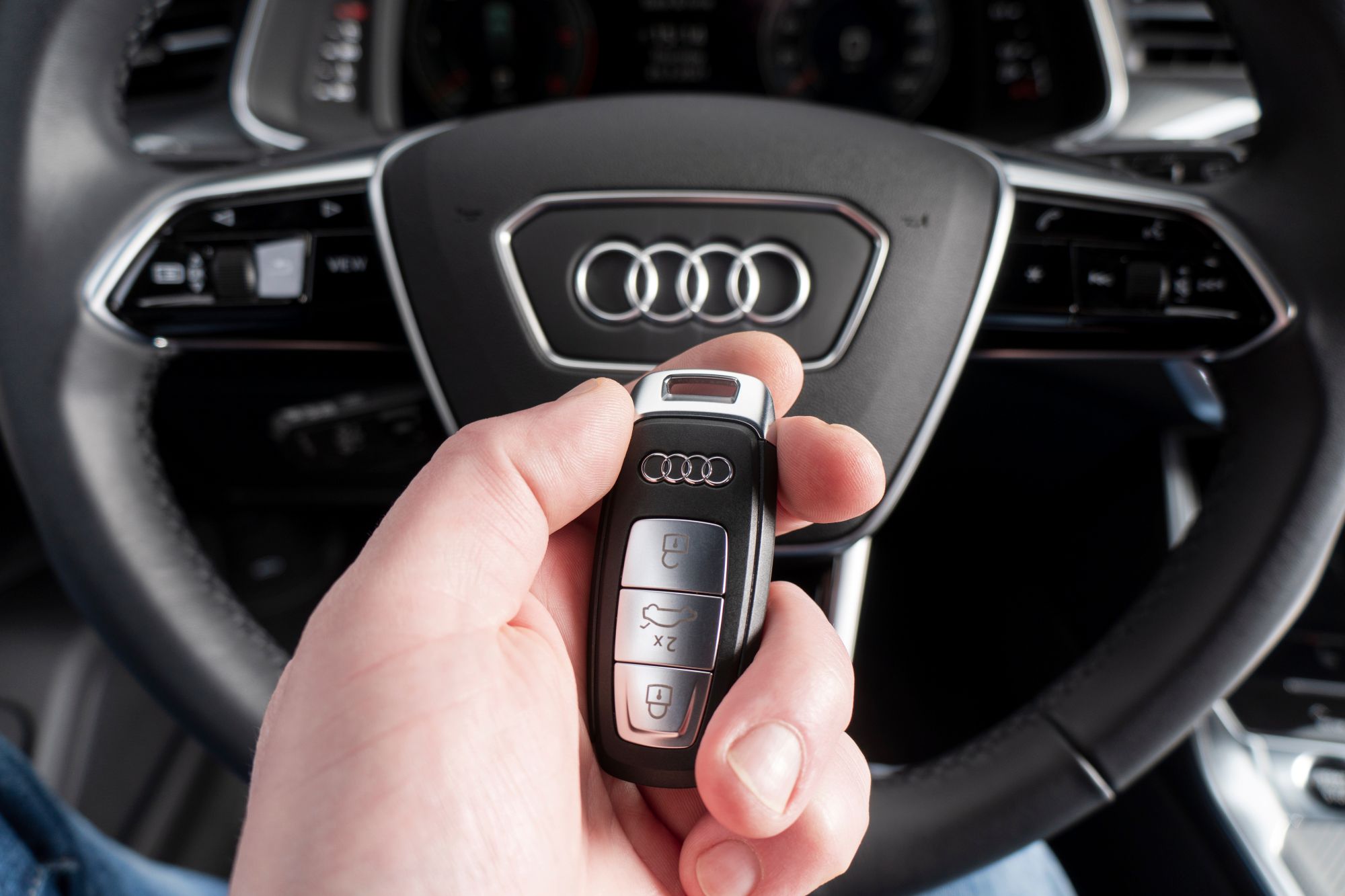 Hand holding an Audi key fob with three silver buttons in front of the steering wheel and dashboard