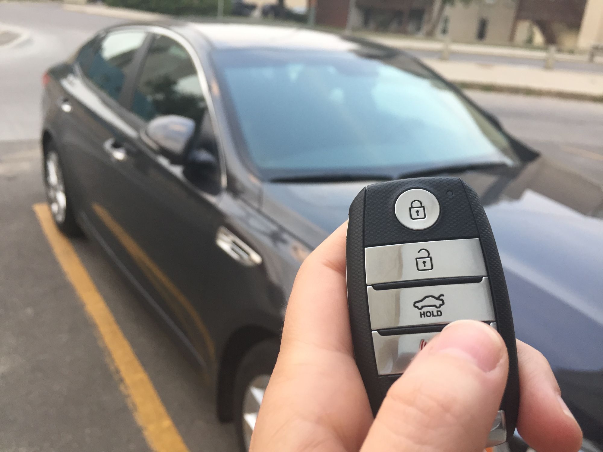 Hand holding a key fob in front of a parked dark sedan
