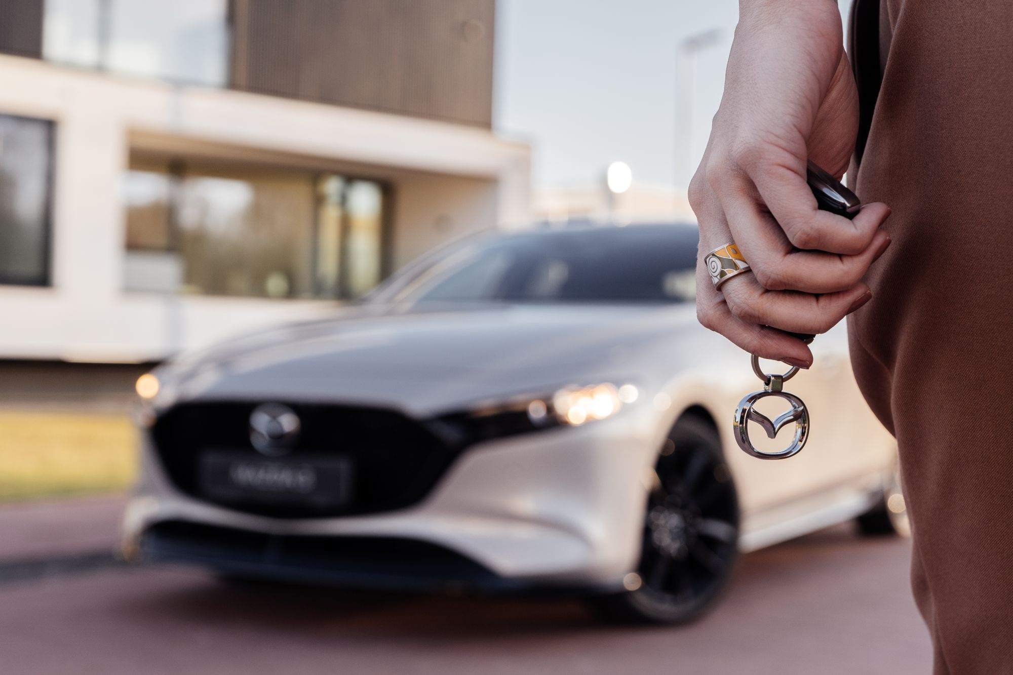 Hand holding a Mazda key fob with a Mazda logo keychain in front of a white Mazda car