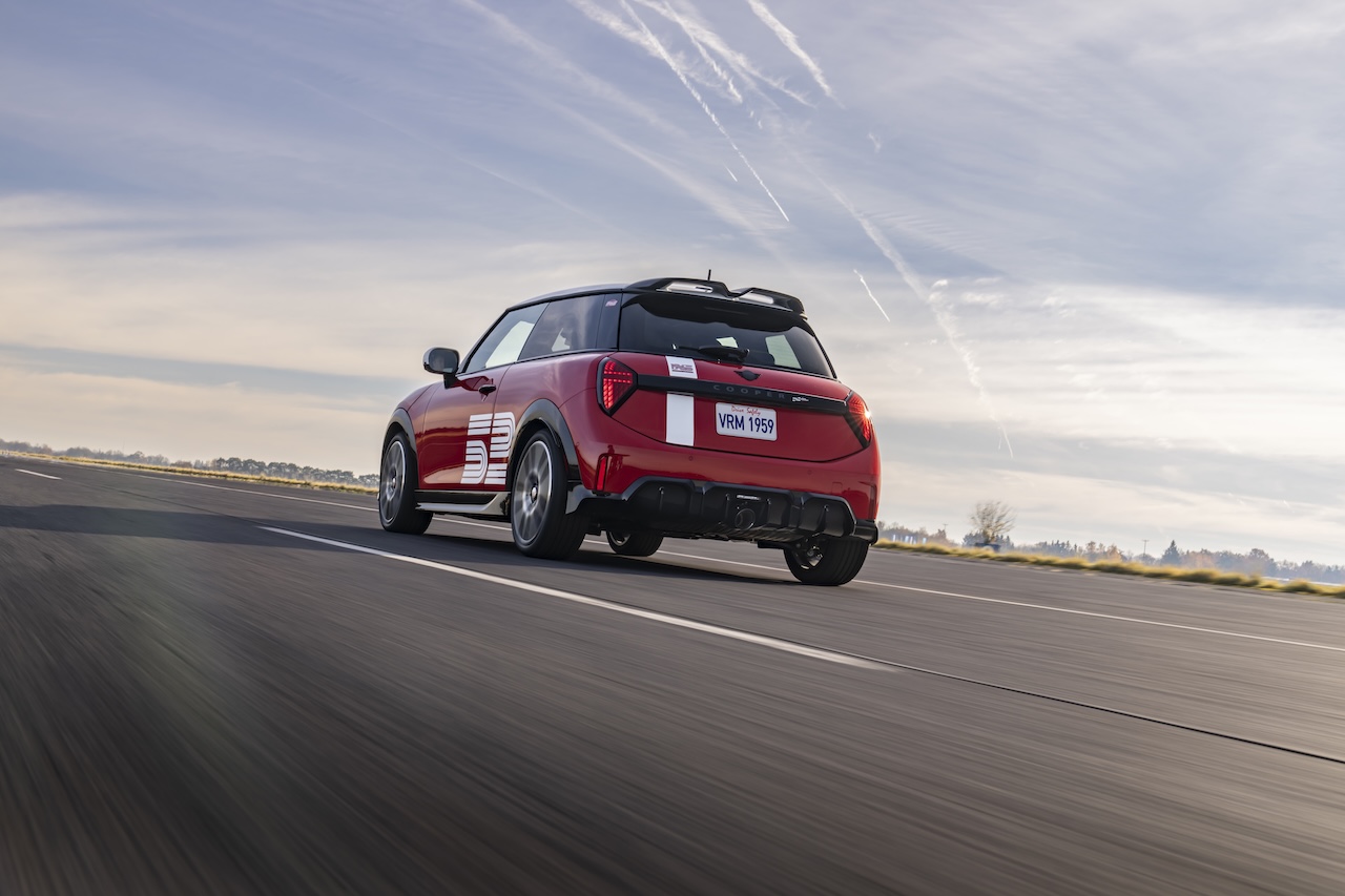 2026 MINI 1965 Victory Edition JCW driving away, rear three-quarter view showing white roof and sport stripes